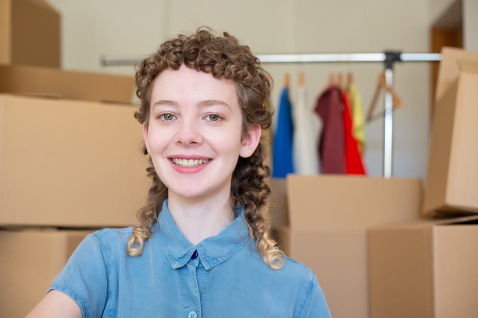 A young woman with curly light brown hair, wearing a light blue collared shirt, smiling at the camera inside a room filled with packed cardboard boxes. Behind her, there is a clothing rack with variou