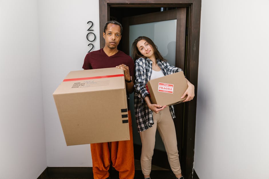 A man and a woman standing in a doorway inside a home, both holding cardboard boxes used for packing and moving. The man, dressed in a burgundy t-shirt and orange trousers, appears to be preparing to load or transport a large cardboard box with packing tape and a label, while the woman, wearing a checked shirt and beige pants, is holding a smaller box with a 'Fragile' sticker, indicating careful handling. Behind them, a dark wooden door frames the entrance to an interior space, and the surrounding walls are painted white. The scene suggests an active home relocation or furniture transport process, with packing materials, boxes, and the presence of a moving or removals service, as exemplified by [COMPANY_NAME], facilitating the packing and loading stages of the moving process. The lighting is bright and natural, capturing the moment of coordinated effort during house removals or packing and moving tasks.