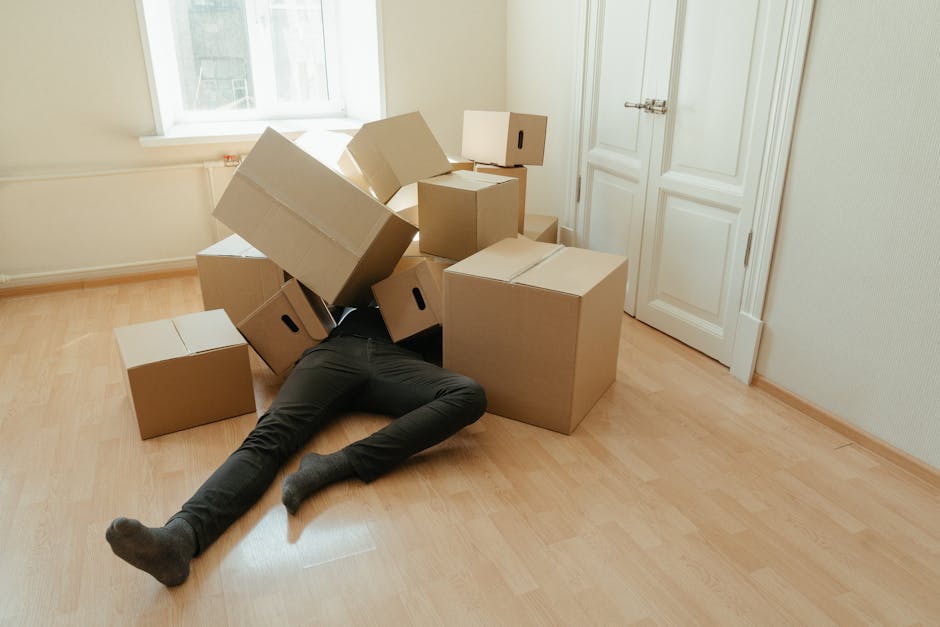 A person dressed in dark pants and socks is lying on a light wooden floor inside a room, partially obscured beneath a stack of cardboard moving boxes of various sizes. Some boxes are open, while others are closed, with one box placed on top of the person’s legs. The room features white walls, a window on the left allowing natural light to brighten the space, and a closed white door with a silver handle on the right side. The boxes appear to be packed for home relocation, reflecting a packing and moving process, with some boxes resting on the floor and others stacked haphazardly. Visible packaging materials include plain cardboard boxes and a slight hint of protective padding or plastic wrap around the boxes. The scene captures the typical activity of furniture transport preparations, with nearby equipment, such as moving trolleys or straps, not visible, but the context suggests professional removals by Man With a Van Lee, specialising in removals services.