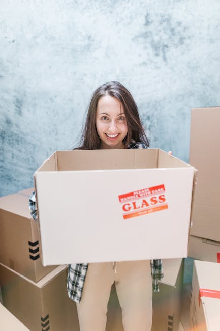 A man and a woman standing in a doorway inside a home, both holding cardboard boxes used for packing and moving. The man, dressed in a burgundy t-shirt and orange trousers, appears to be preparing to load or transport a large cardboard box with packing tape and a label, while the woman, wearing a checked shirt and beige pants, is holding a smaller box with a 'Fragile' sticker, indicating careful handling. Behind them, a dark wooden door frames the entrance to an interior space, and the surrounding walls are painted white. The scene suggests an active home relocation or furniture transport process, with packing materials, boxes, and the presence of a moving or removals service, as exemplified by [COMPANY_NAME], facilitating the packing and loading stages of the moving process. The lighting is bright and natural, capturing the moment of coordinated effort during house removals or packing and moving tasks.