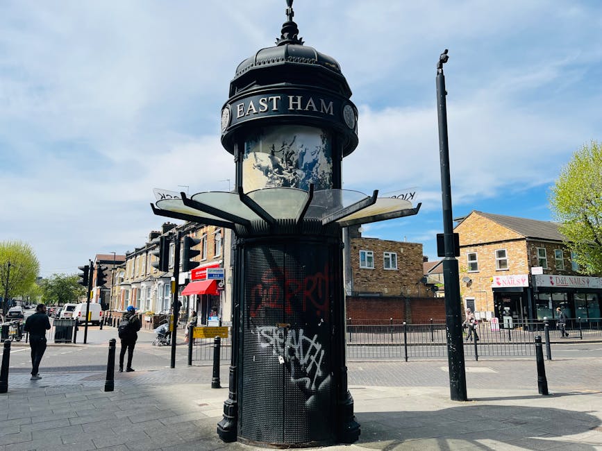 A vintage-style black and white cylindrical public notice board with a domed top featuring the sign 'EAST HAM' on a busy street corner. The structure includes a central pole supporting a glass enclosure for notices, and the base is covered with black metal panels, some of which are vandalized with graffiti. Surrounding the notice board are traffic lights, pedestrian crossing signals, and several pedestrians walking across the pavement. In the background, there are brick residential buildings and retail shops, including a nail salon, with green trees lining the street under a partly cloudy sky. The scene captures a typical urban environment, relevant to home relocation and moving logistics in the context of house removals or furniture transport, as seen in the professional services provided by companies like Man With a Van Lee.