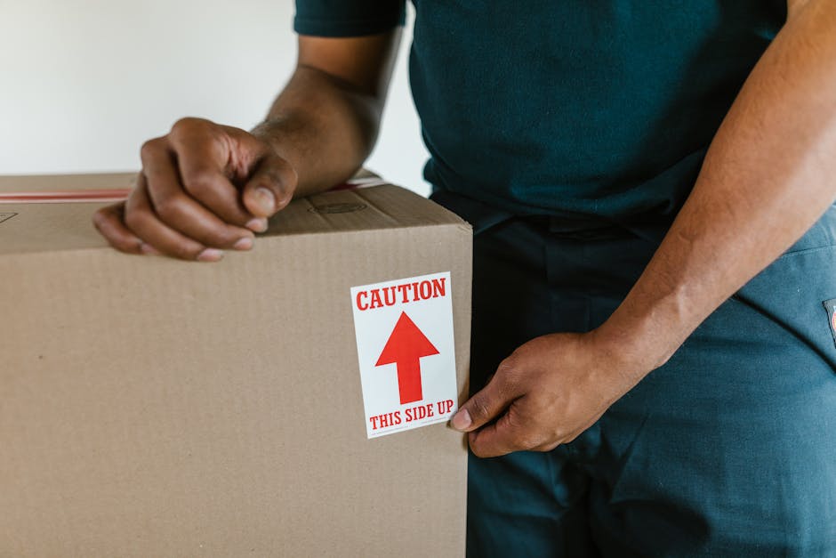 Close-up of a person wearing a blue t-shirt and dark trousers, carefully gripping a large cardboard box sealed with brown packing tape. The box features a red and white safety label prominently displaying the word 'CAUTION,' accompanied by an upward-pointing arrow and the instruction 'THIS SIDE UP,' indicating proper handling during furniture transport and home relocation. The individual is positioned indoors, possibly in a hallway or loading area, with a neutral background and natural lighting. The scene emphasizes the safe packing practices involved in house removals and the meticulous approach of a professional removals service such as Man With a Van Lee, who handles packing, furniture transport, and moving logistics with care during the loading process for office or home relocations.
