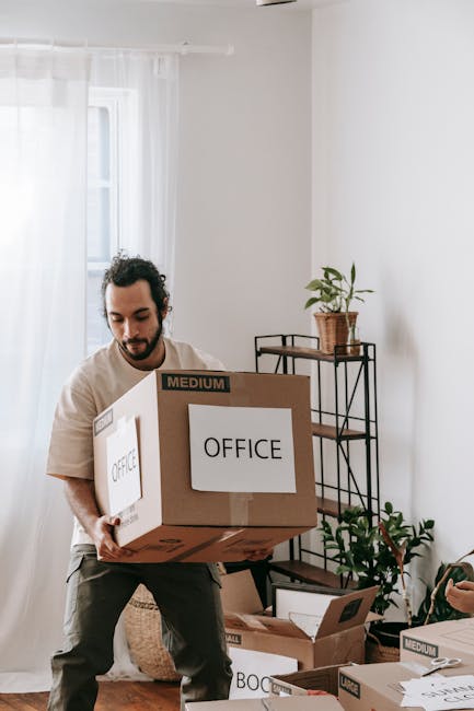 Close-up of a person wearing a blue t-shirt and dark trousers, carefully gripping a large cardboard box sealed with brown packing tape. The box features a red and white safety label prominently displaying the word 'CAUTION,' accompanied by an upward-pointing arrow and the instruction 'THIS SIDE UP,' indicating proper handling during furniture transport and home relocation. The individual is positioned indoors, possibly in a hallway or loading area, with a neutral background and natural lighting. The scene emphasizes the safe packing practices involved in house removals and the meticulous approach of a professional removals service such as Man With a Van Lee, who handles packing, furniture transport, and moving logistics with care during the loading process for office or home relocations.