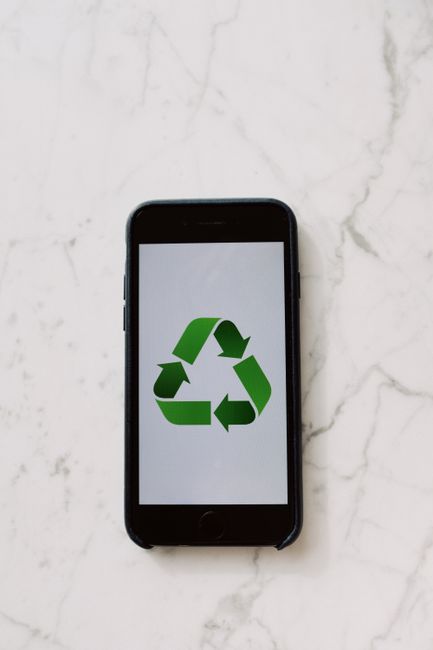 A black smartphone lying flat on a light-colored marble surface, displaying a prominent green recycling symbol composed of three arrows forming a triangle. The phone is centered in the image, with its screen occupying most of the frame, and the recycling icon clearly visible. The marble surface has subtle gray veining and provides a neutral background. This image could relate to environmentally conscious practices involved in house removals and relocation services, such as recycling packing materials or promoting eco-friendly moving methods, as part of the offerings by Man With a Van Lee. The setting appears to be indoors, possibly in a home or office environment, and the lighting is soft and even, highlighting the phone and the recycling symbol on its screen.