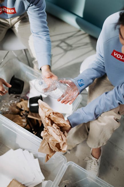 Two individuals wearing light blue uniforms with red 'VOLUNTEER' patches are engaged in a packing process indoors, preparing items for a house move or relocation. One person is holding a clear plastic bottle while the other is wrapping it with bubble wrap, focusing on fragile object protection. They are working over an open plastic storage container filled with crumpled brown paper and packing materials, indicating careful packing and handling of household items. There are additional packing supplies, such as cardboard boxes and white papers, scattered around on the floor and inside the container. The setting appears to be a modern interior space with soft lighting and a light-colored floor, suggesting the early stages of a home removal or furniture transport process. The activity is part of a professional removals or moving service, possibly operated by Man With a Van Lee, focused on environmentally conscious packing, reuse, and recycling of packing materials.