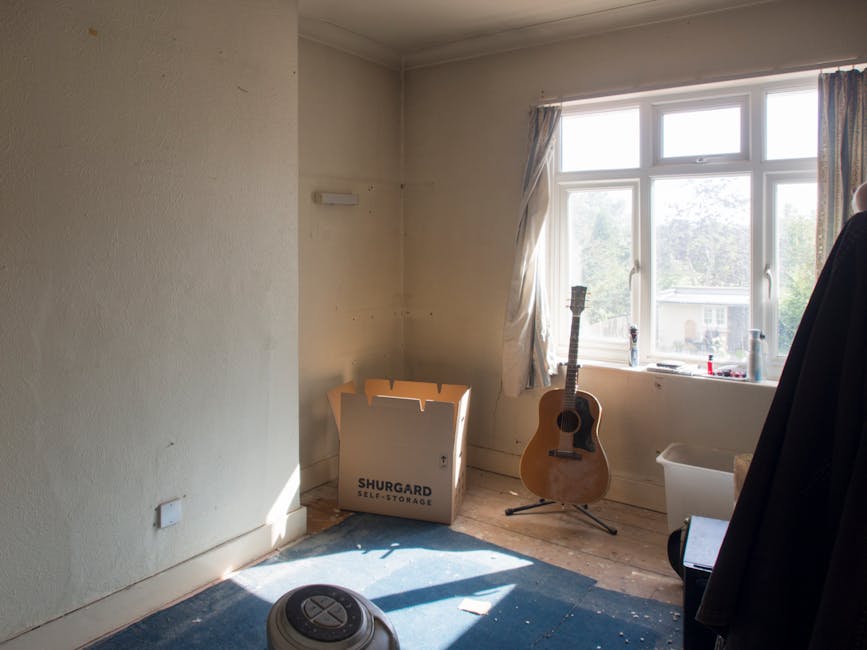 Inside a sparsely furnished room with cream-colored walls and a large window allowing natural daylight to illuminate the space, a cardboard box labeled 'SHURGARD SELF-STORAGE' is positioned near the corner, indicating recent packing or move preparations. Adjacent to the box, a wooden acoustic guitar on a stand is placed upright, suggesting personal items ready for transportation. The window reveals a view of neighboring houses and greenery, with curtains pulled to the sides. On the floor, a partially visible vacuum or cleaning device rests on a dark carpet or mat, and next to the window, a white plastic storage bin and a black jacket hanging on a chair or hook are observable. The scene captures a moment in the home relocation process, illustrating the packing stage before or after a move, consistent with the services offered by Man With a Van Lee for end of tenancy removals, as indicated in the page title. The environment appears tidy but in the midst of an ongoing packing or moving operation.