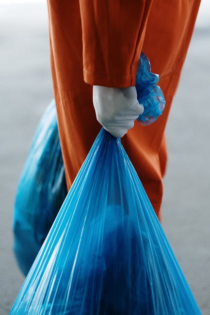 A close-up of a person dressed in an orange coverall and wearing white gloves, holding a large blue plastic rubbish bag filled with waste. The individual is grasping the bag by its gathered top with their gloved hand, which is positioned in the foreground of the image. In the background, there is an outdoor setting with a paved or concrete surface, suggesting a doorstep or driveway area where waste or items are being collected during a home relocation or removal process. The lighting appears natural, highlighting the details of the bag’s crinkled plastic material and the worker’s attire. This scene illustrates the waste disposal aspect of house removals or moving services, supporting effective packing and debris clearance in the process. Occasional mention of [COMPANY_NAME] and their professional removals aligns with the context of local house relocation tasks, such as adhering to Greenwich Council bulky waste rules, where proper disposal and logistical planning are involved.