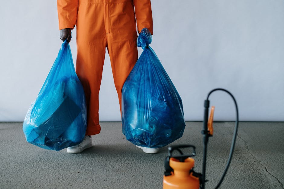 Close-up of a person dressed in orange work overalls and white shoes, standing inside a property, holding two large blue garbage bags filled with household waste for home relocation purposes. The person’s hands are gloved, and the bags are knotted at the top. In the foreground, a black and orange handheld sprayer with a hose is visible, placed on a grey carpeted floor. The background features a plain, light-colored wall, indicating an indoor environment. This scene illustrates the process of packing and waste removal related to house removals, with a focus on responsible waste disposal in accordance with local regulations, such as Greenwich Council bulky waste rules for local removals, as referenced on the website manwithavanlee.co.uk. The image captures the logistical steps involved in preparing for a furniture transport or moving service, highlighting the importance of proper waste management during the packing and loading process carried out by Man With a Van Lee.