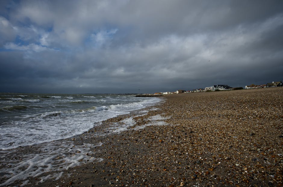 A wide view of a pebble beach with scattered small stones and shells, leading to the shoreline where ocean waves are gently washing onto the sand, under a cloudy sky with dark grey clouds and some patches of lighter sky. In the distance, a row of houses and buildings is visible along the coastline, with some structures having pitched roofs. The environment suggests an overcast day, typical for coastal weather, with diffused natural lighting. This scene illustrates a seaside setting, potentially involved in house removals or relocating services near the coast, although no people or removal equipment are visible. The image emphasizes the landscape and open space characteristic of seaside locations, suitable for outdoor logistics or packing and moving preparations near a coastal home, as managed by [COMPANY_NAME].