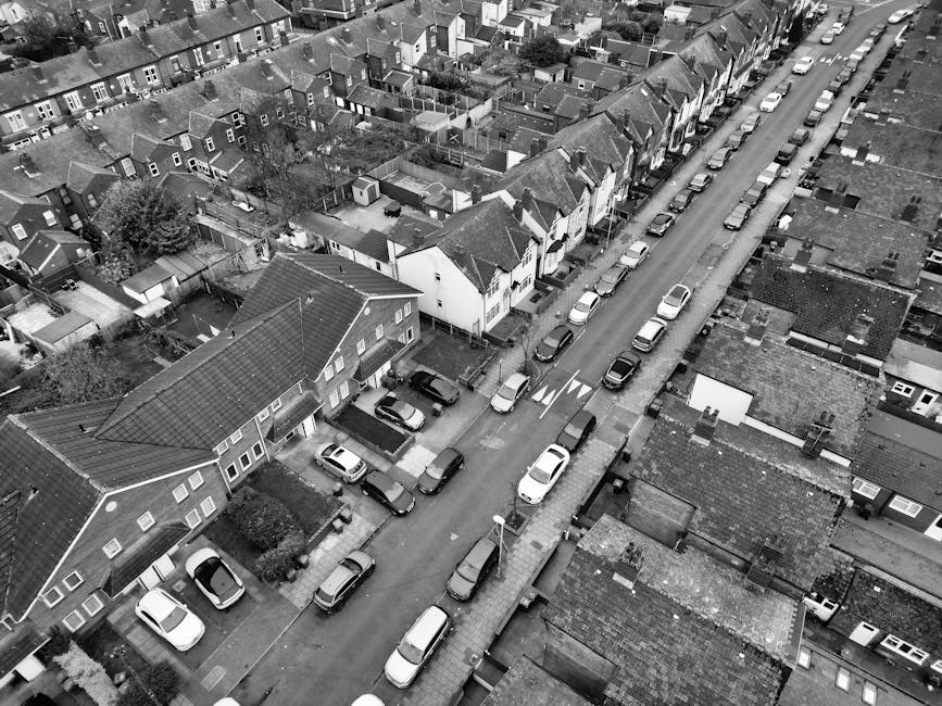 An aerial black-and-white photograph of a residential street showing rows of terraced houses with pitched roofs, front gardens, and driveways. Several cars are parked along both sides of the narrow road, which includes a section with painted directional arrows indicating traffic flow. The street features a concrete sidewalk with a street lamp visible near the centre of the image. In the foreground, part of a house with a tiled roof and a small front yard is visible, while behind it, a driveway with a vehicle parked on it is seen. The surroundings include closely packed houses with small backyards, sheds, and trees, indicative of a typical suburban neighbourhood. The image captures the environment in daylight, providing a detailed view of the housing and parking arrangement typical for home relocation logistics, with a focus on tight urban access challenges managed by professional removals facilitated by companies like Man With a Van Lee.