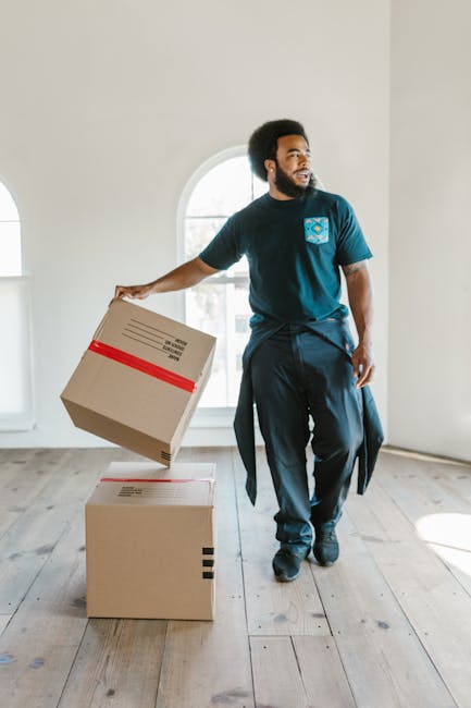 A man with an afro hairstyle and a beard, dressed in a navy blue t-shirt with a printed logo on the chest and dark trousers, stands inside a bright, empty room with high ceilings and a large arched window. He is holding a cardboard box with red packing tape around it, preparing for a home relocation. In front of him, two other cardboard boxes are placed on the light wooden floor, one on top of the other, indicating a loading or packing process typical of furniture transport and moving logistics. The scene is well-lit with natural daylight streaming through the window, emphasizing the indoor environment associated with house removals. Man With a Van Lee specializes in removals, and this image illustrates the packing stage of an organized house move involving careful handling of boxed belongings and efficient loading procedures.