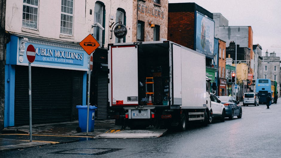 A white moving van with an open rear door is parked on the street outside a commercial building, ready for a home relocation or furniture transport process. Inside the van, visible storage shelves hold packing supplies such as plastic containers and cardboard boxes, indicating an organized packing and moving operation. The van is positioned partly on the pavement with its loading ramp extended onto the street, suggesting the unloading or loading of household items. Nearby, a blue recycling bin is situated next to a no-entry traffic sign, and a street sign depicts a restricted vehicle zone ahead. The surrounding area features a row of shops and businesses with various signage, including a blue storefront for Dublin Mouldings, and other commercial establishments with signage and advertisements advertising local services. The street is wet, and the ambient light suggests daytime, with some vehicles and pedestrians visible in the background, emphasizing the urban environment typical for professional removals and transport services by companies like Man With a Van Lee.