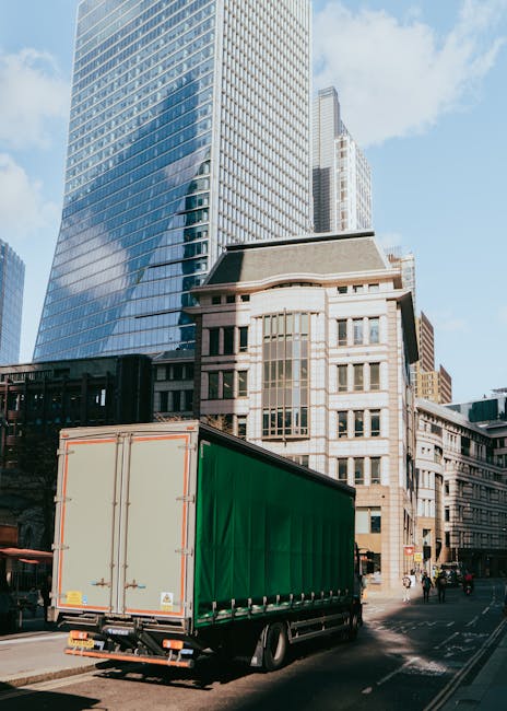 A large commercial truck with a green cargo area is parked on an urban street, surrounded by tall modern office buildings and older architectural structures. The truck is positioned close to the curb,