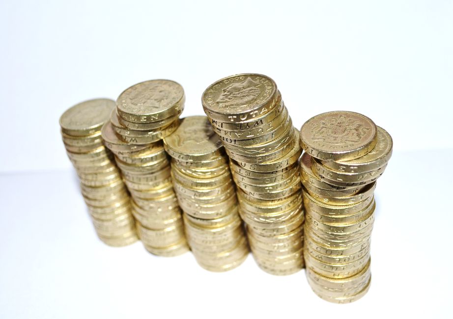 Stacked British one-pound coins arranged in five vertical columns on a white surface, with some coins slightly tipped or overlapping, illustrating money or savings. The background is plain and bright,