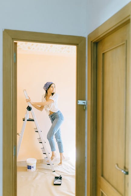 A woman wearing a headscarf, white blouse, and light blue jeans is standing on a step ladder inside a room, engaged in a home relocation activity. She is holding a paintbrush or small roller and appears to be preparing the walls for painting or decorating as part of the moving process. The room has light-colored walls and a bright, well-lit environment. On the floor, there is a paint tray and a container of paint, indicating ongoing decorating work. The image is viewed through a wooden doorway, and the perspective suggests it is part of a house removal or refurbishment project coordinated by Man With a Van Lee, a professional moving company. The scene captures the lighter, preparatory stages of house removals and furniture transport, emphasizing packing, decorating, and logistical arrangements involved in house relocations.