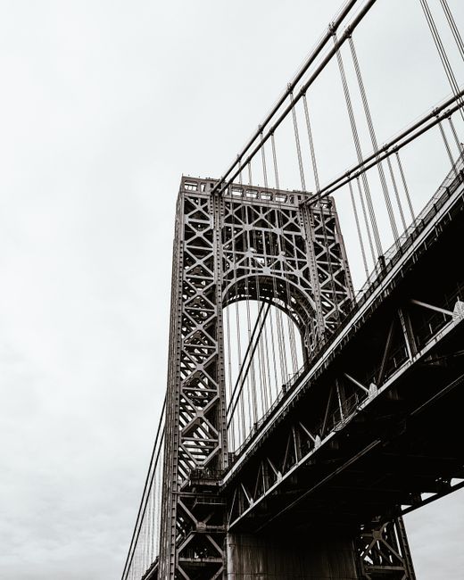 A black-and-white photograph of the upper section of the Brooklyn Bridge showing its iconic steel tower with crisscrossing beams and vertical cables. Part of the bridge's roadway structure is visible beneath, along with suspension cables extending diagonally upward from the tower. The sky in the background is overcast, creating a neutral backdrop. This image captures the architectural details of the bridge, which is often associated with city relocation and transport infrastructure, relevant to the context of house removals and moving services provided by Man With a Van Lee. The focus is on the structural elements that support the bridge, emphasizing its engineering design and historical significance in urban moving logistics.